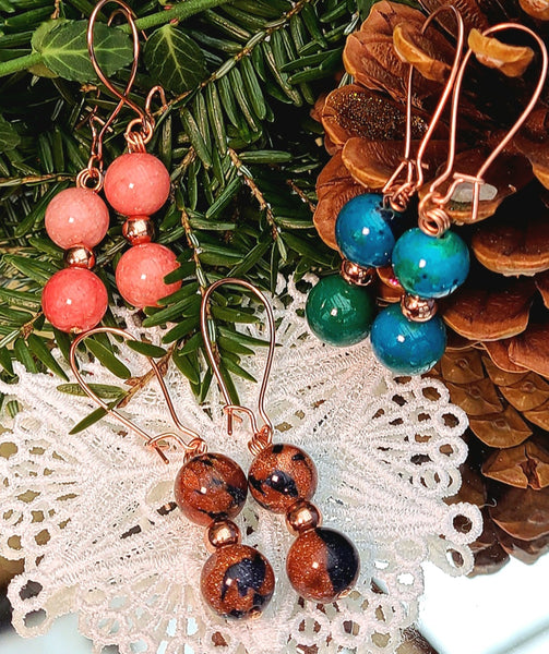 Image of earrings with two 10mm beads; one pair Sandstone, one pair Phoenix and one pair Pale Red Chalcedony,  each separated by a 5mm copper bead in a display with a pinecone, pine branches and a white artificial snowflake