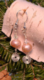 Image of a pair of earrings with a 10mm freshwater pearl topped with a clear crystal and a circular spiral of silver wire hanging below; on birch bark in front of pine braches