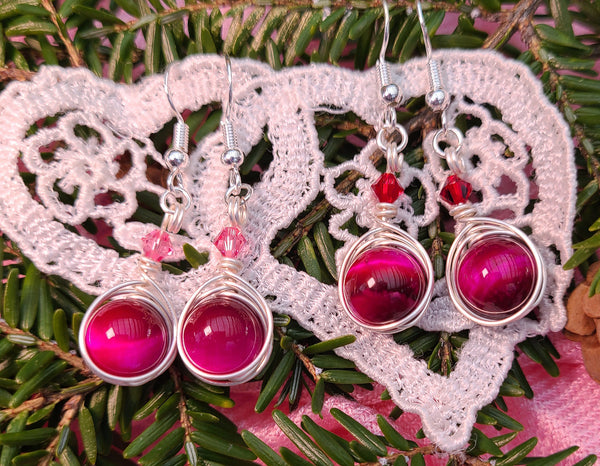 Image of 2 pairs of earrings with pink tiger eye 10mm beads wrapped by silver wire and topped by a red or pink 4mm Swarovski crystal hanging on a pine branch over white lace hearts