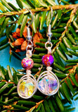 Image of a pair of earrings with 10mm AB coated spiral crystals wrapped with silver wire and topped with galaxy jasper hanging from a pine branch